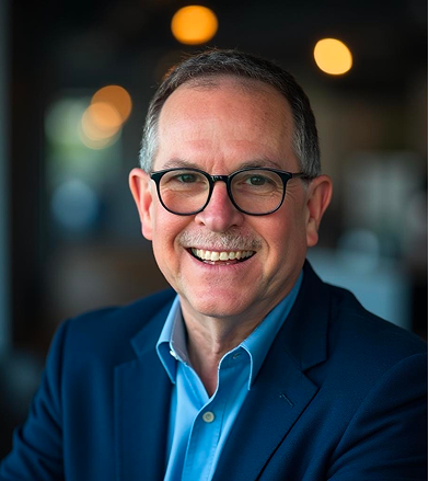 Smiling middle-aged man in glasses and a blue blazer, seated indoors with a softly lit background
