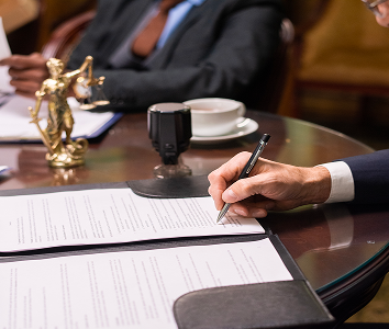 Hand signing legal documents with a justice statue and coffee cup in the background.